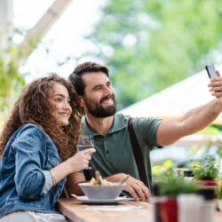 Happy couple sitting outdoors on terrace restaurant, taking selfie