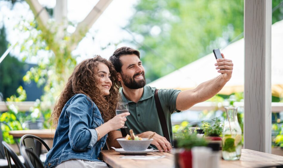 Happy couple sitting outdoors on terrace restaurant, taking selfie